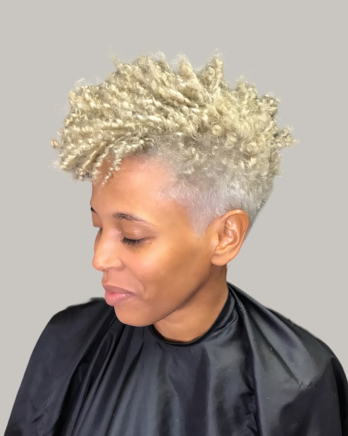 Woman in a salon cape with a platinum blonde tapered afro, looking down