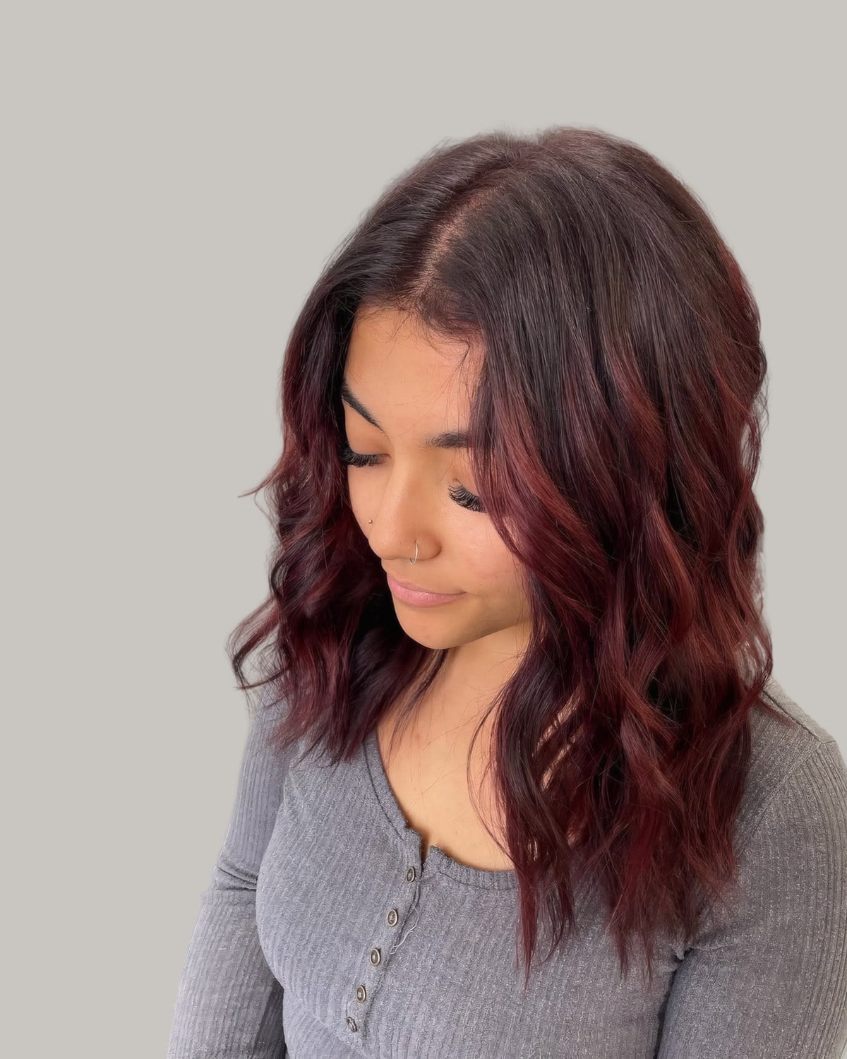Woman looking down with a dark root and plum red waves, in a gray henley