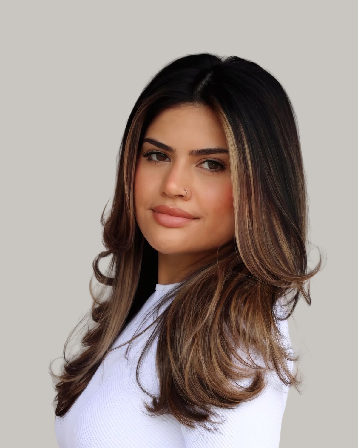 Woman in a white top with a dark root, caramel face-framing pieces and blown-out layers