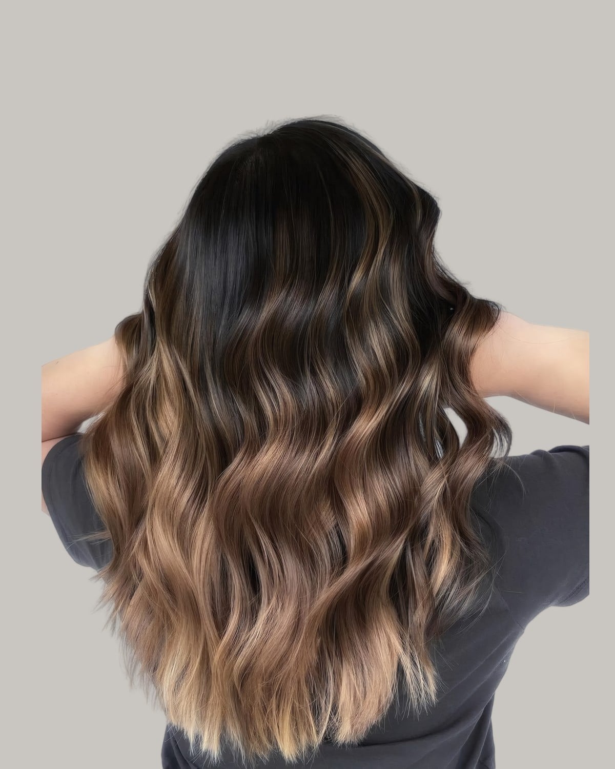 Woman seen from behind, hands in her hair, with a dark root and light brown lengths