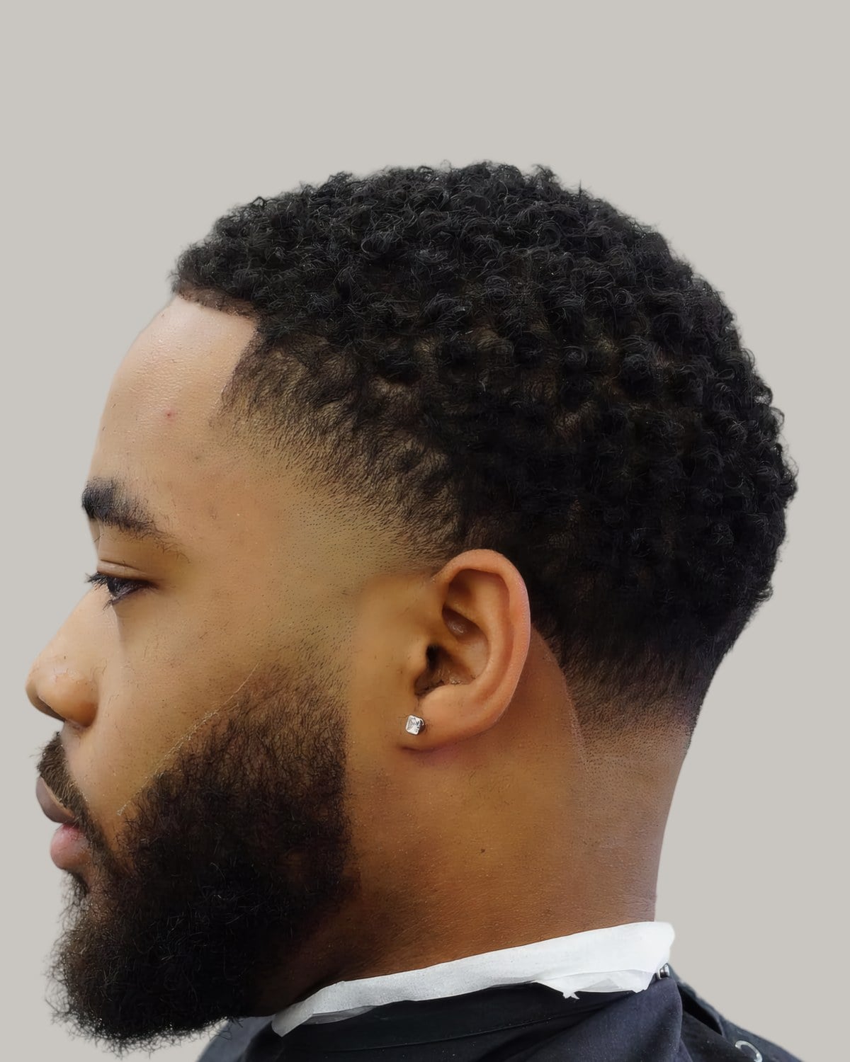 Bearded man with short sponge curls, a stud earring and a drop fade, seen in profile
