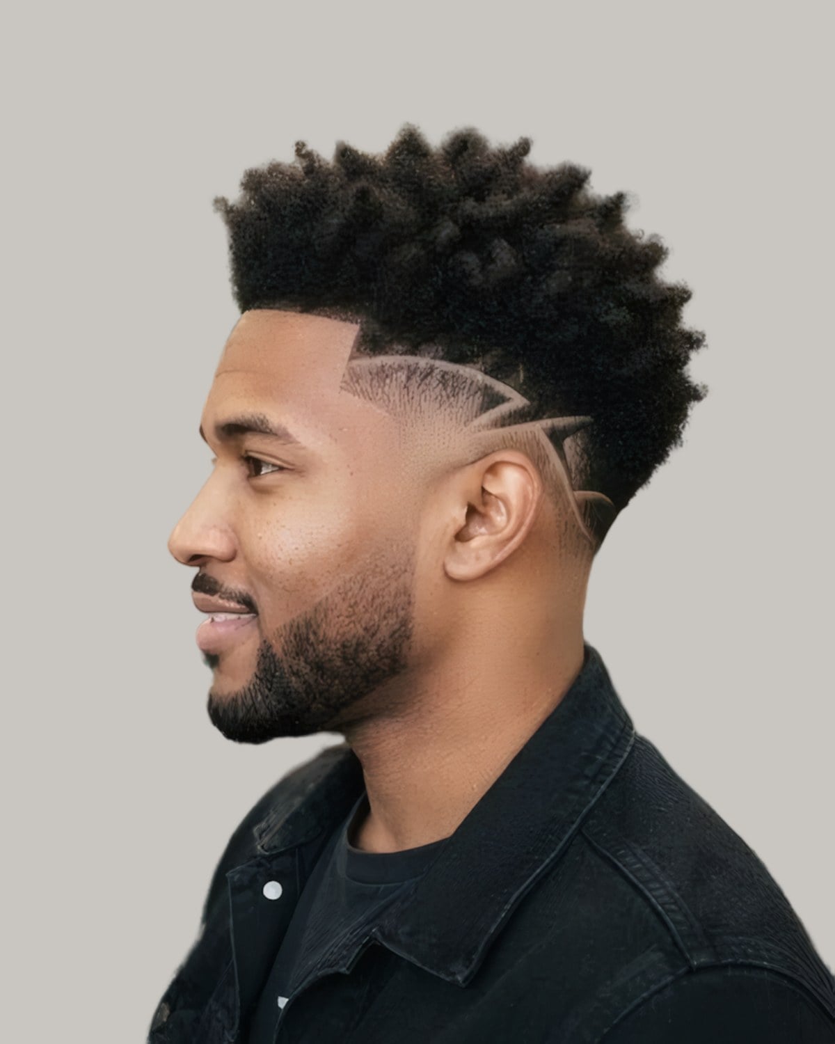 Bearded man with a curly top, a low taper and lines shaved above the ear, seen in profile
