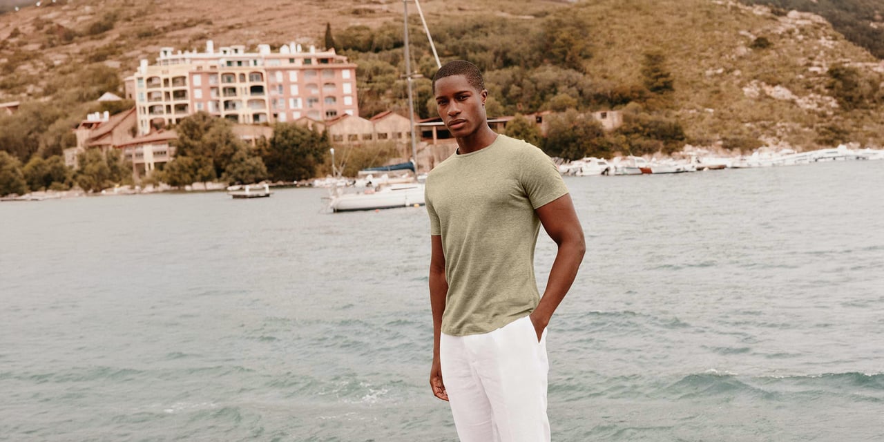 Man with a short low cut in an olive T-shirt and white trousers, standing by the sea with a harbor town behind him