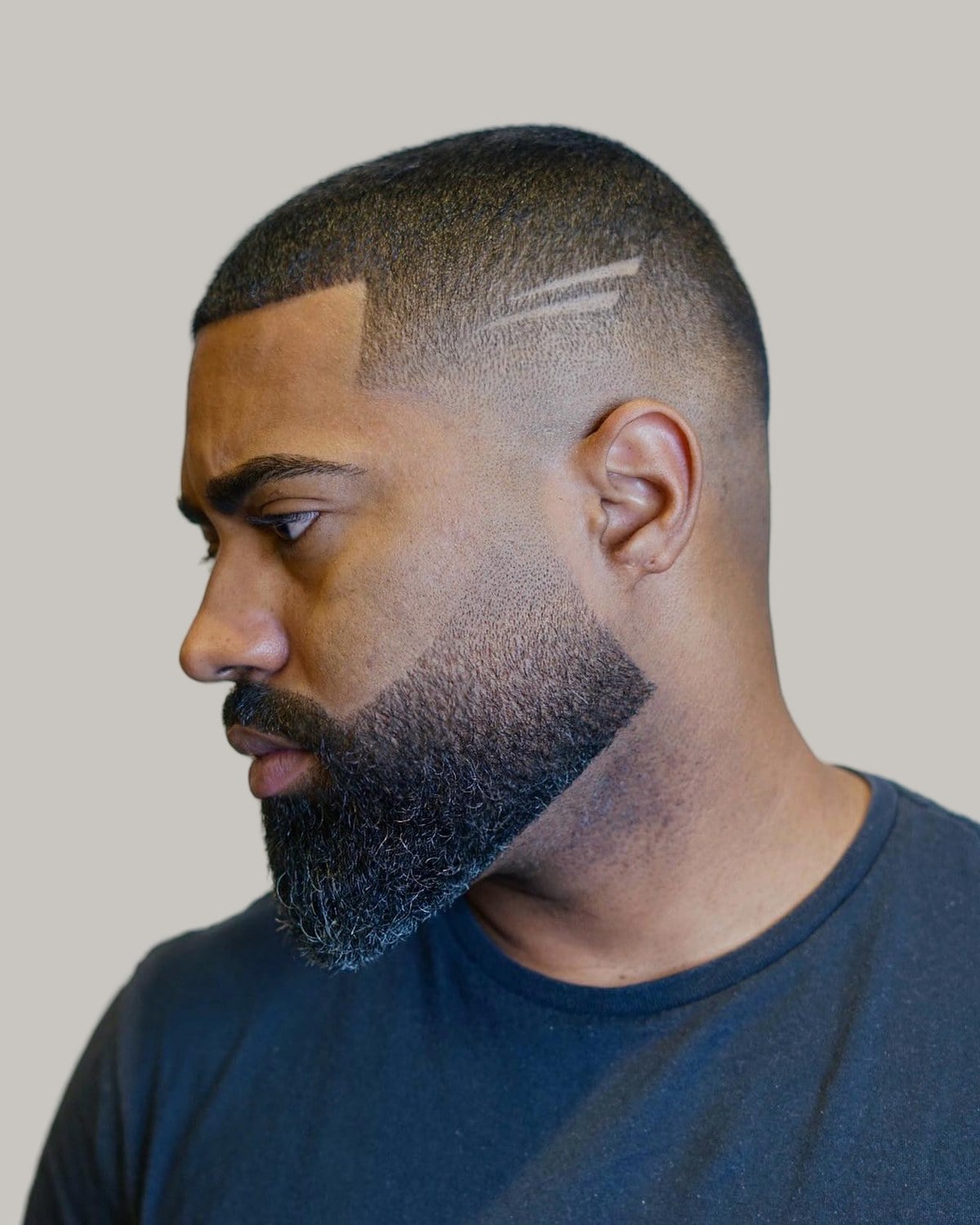 Man with a full beard, a short top and two lines shaved at the temple, seen at an angle