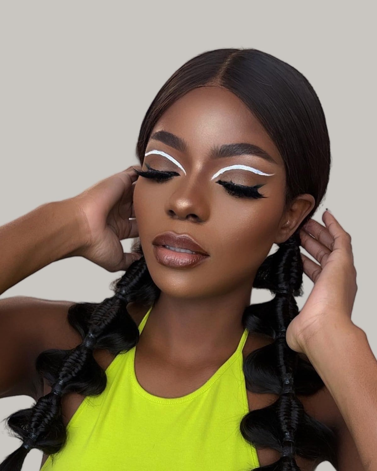 Woman with sleek black bubble pigtails and white graphic eyeliner, her hands at her head