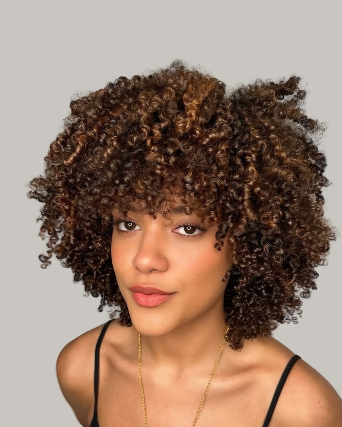 Woman facing the camera with full, rounded curls and a curly fringe, dark at the root with caramel through the lengths