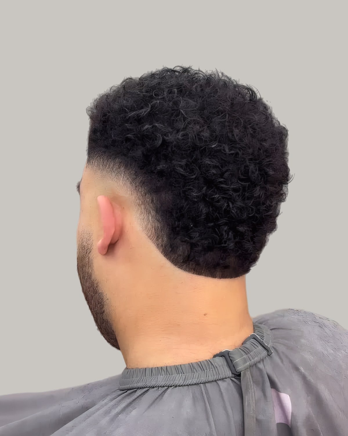 Bearded man with a curly top and a low burst taper, seen from behind at an angle