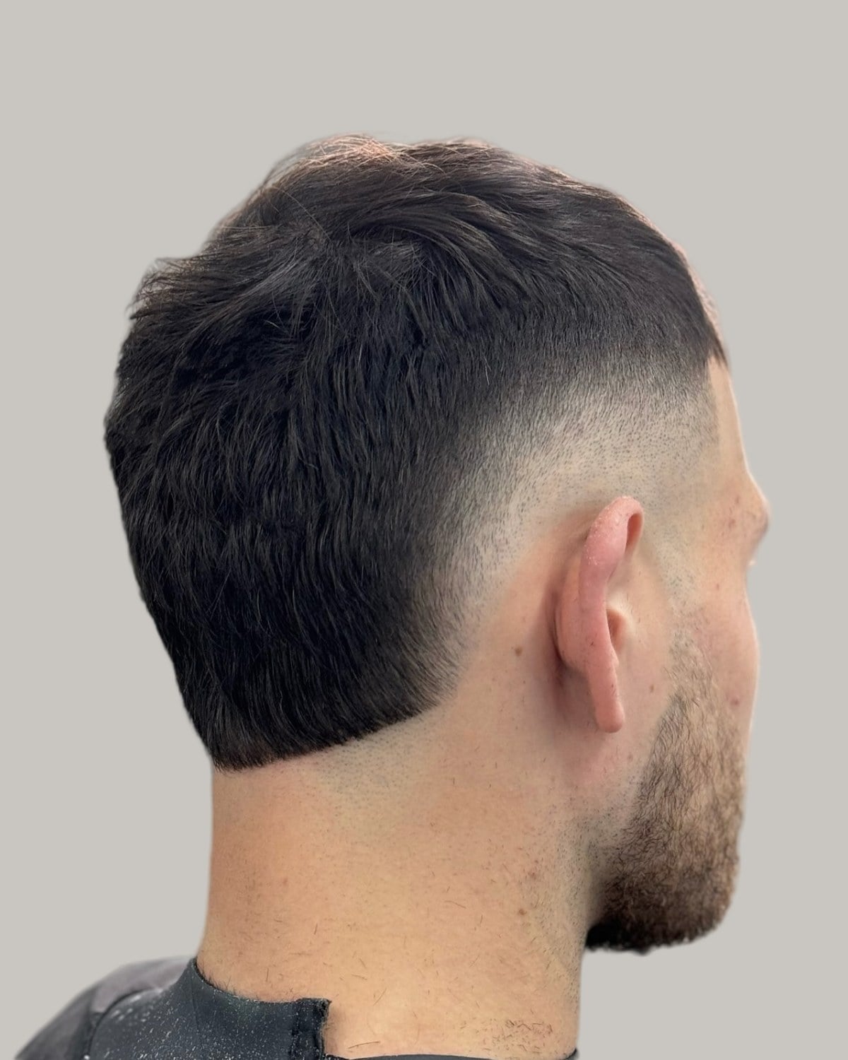 Bearded man with a short dark textured haircut and a low burst fade, seen from behind at an angle