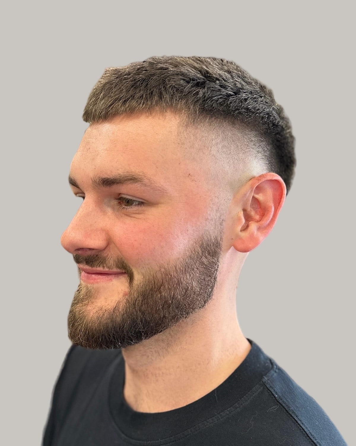 Bearded man with a short buzz cut and a burst fade, smiling and looking to the side