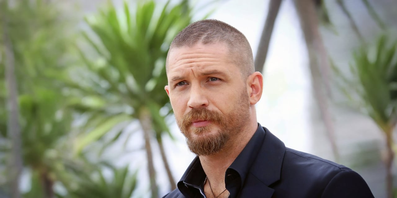 Tom Hardy with a buzz cut and a full beard, in a dark shirt in front of palm trees