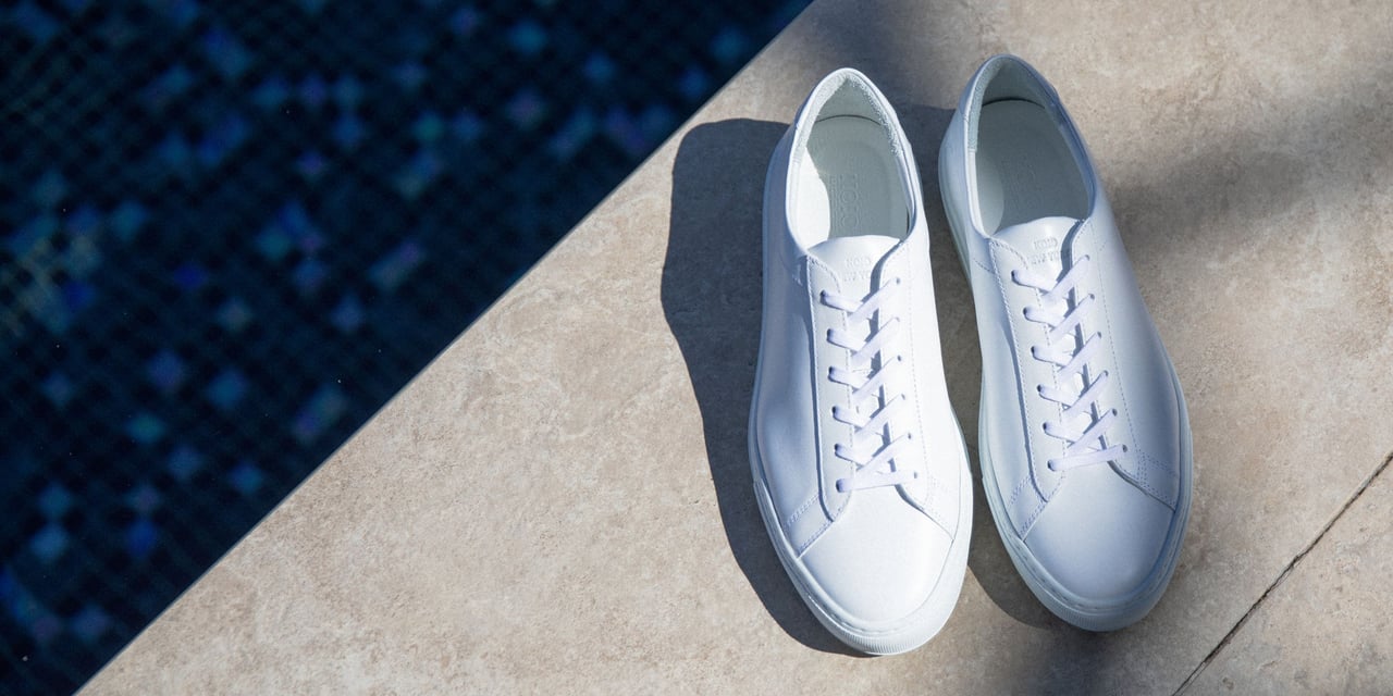 a pair of white leather low-top sneakers on light stone by a pool - Luxe Digital