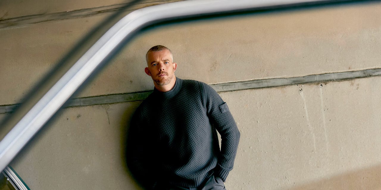 Man with a short crew cut and a trimmed beard, in a dark knit sweater, leaning against a concrete wall
