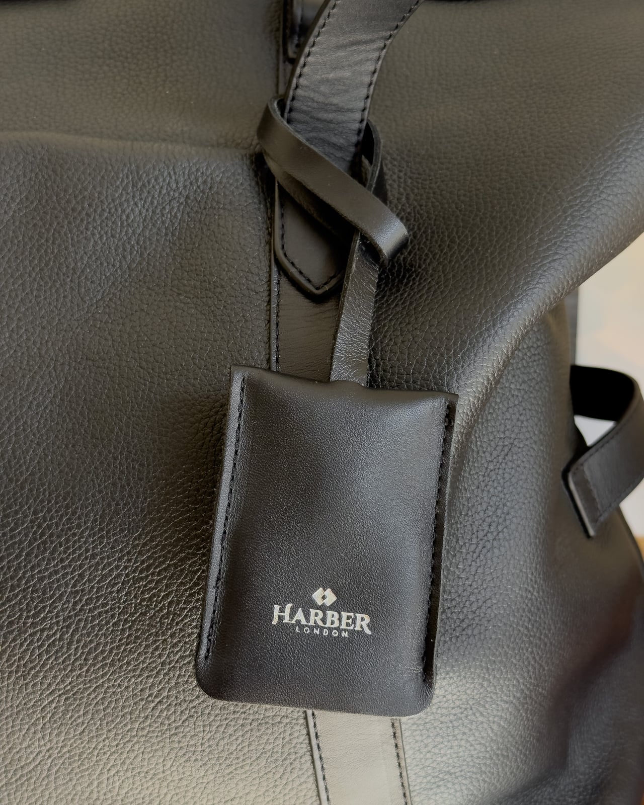 Black leather Harber London AirTag holder hanging from a handle of the Overnight Bag - Luxe Digital