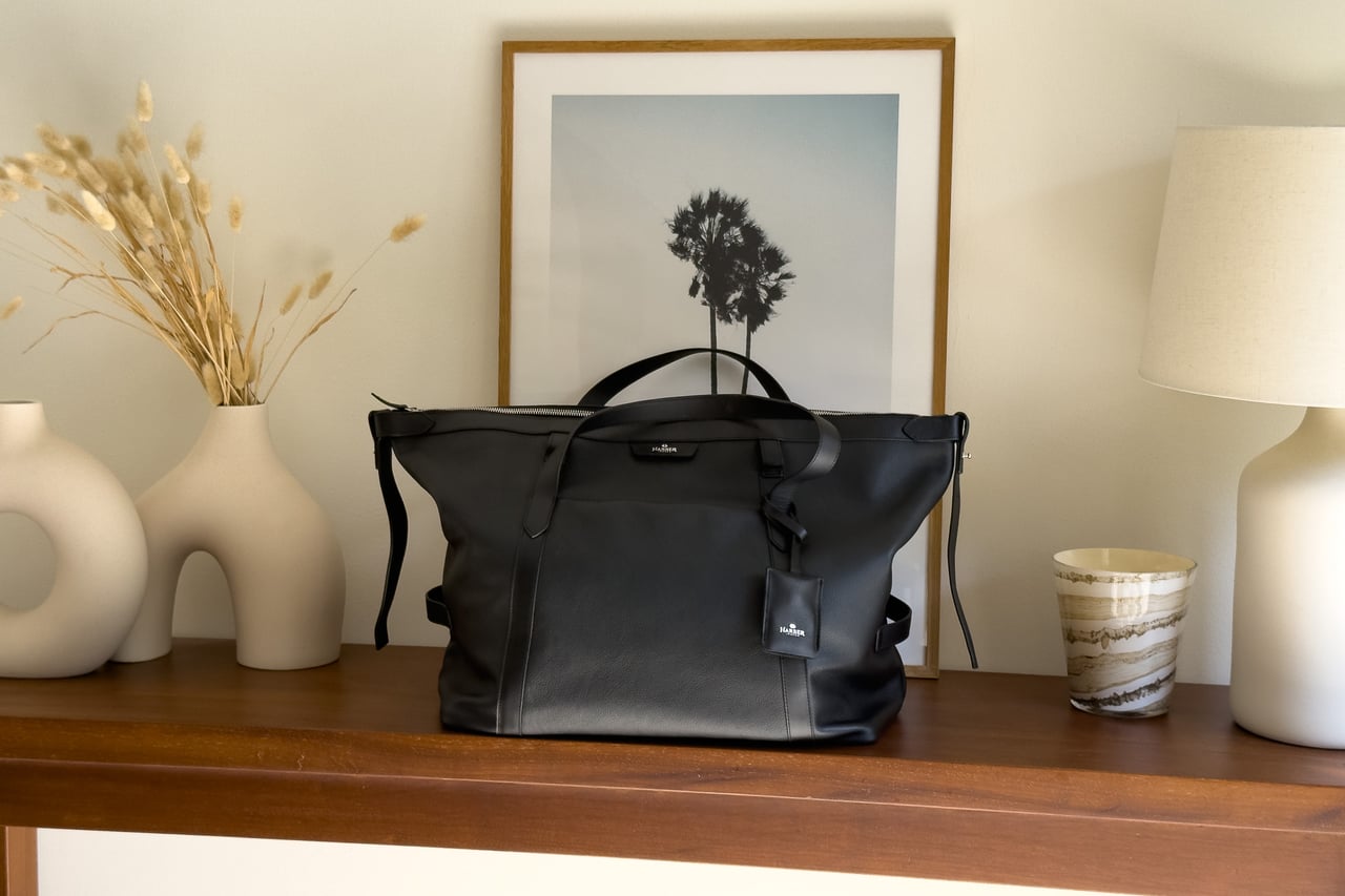 Harber London Leather Overnight Bag in black on a wooden console in front of a framed palm-tree print - Luxe Digital