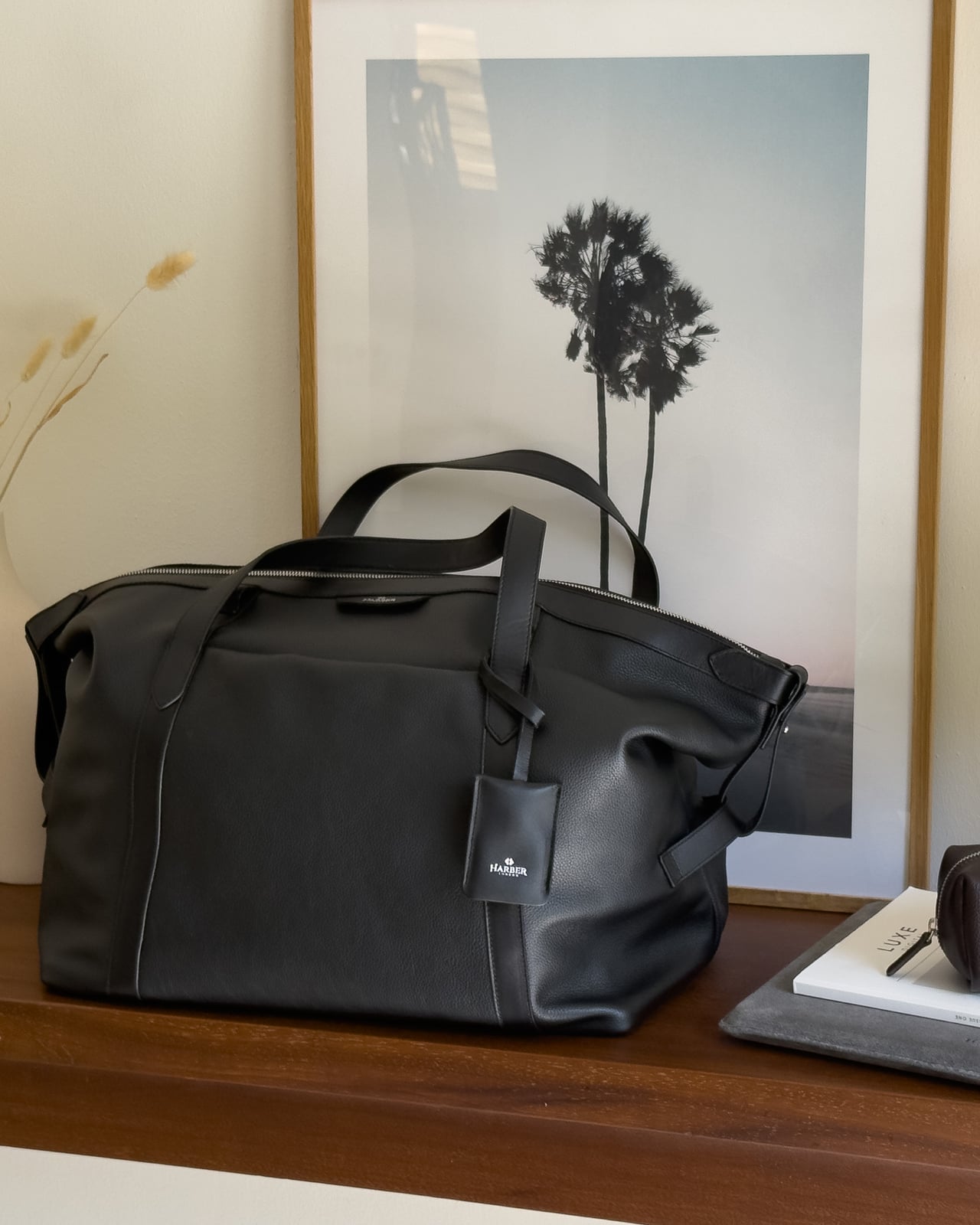 Black Harber London Leather Overnight Bag on a wooden console beside a framed print - Luxe Digital