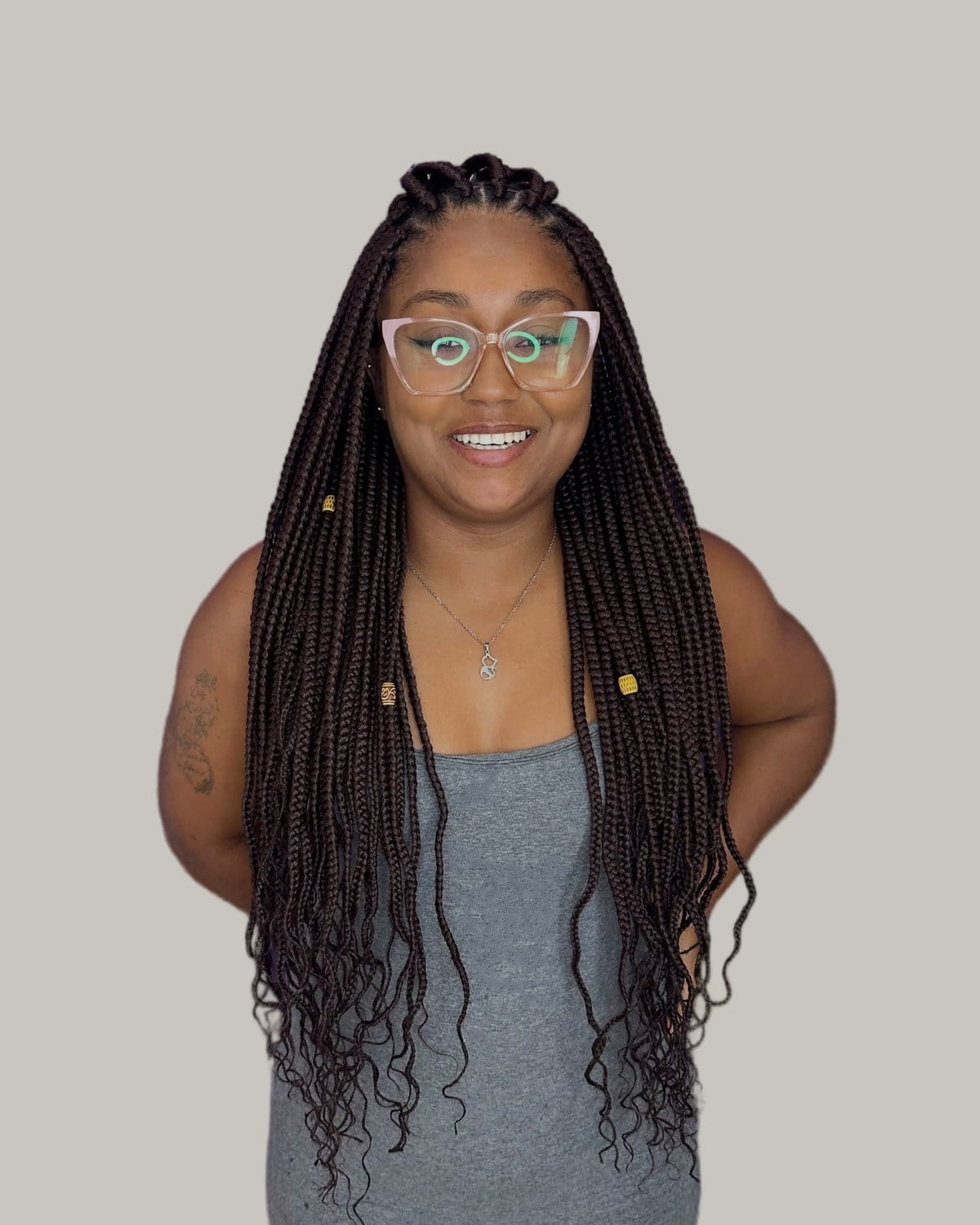 Smiling woman in glasses and a grey tank top with long dark-brown knotless braids and curly ends