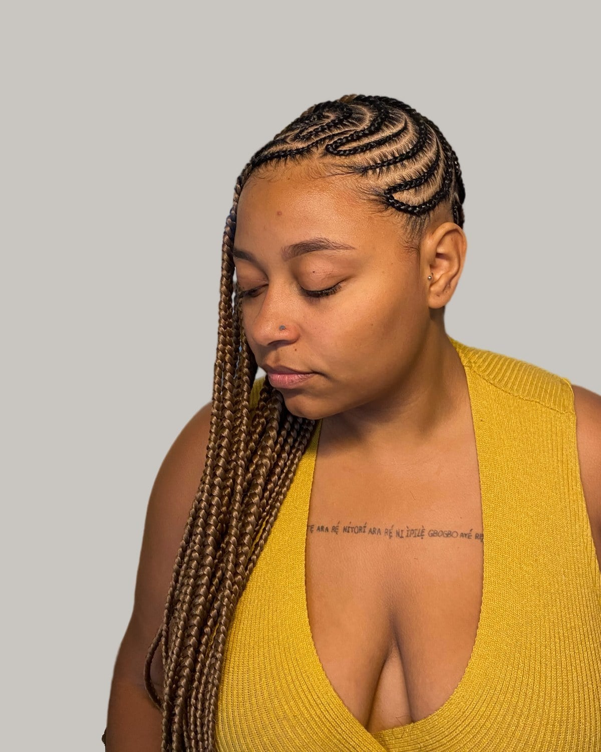Woman in a mustard top looking down, with curved cornrows at the front and long honey-brown braids falling over one shoulder