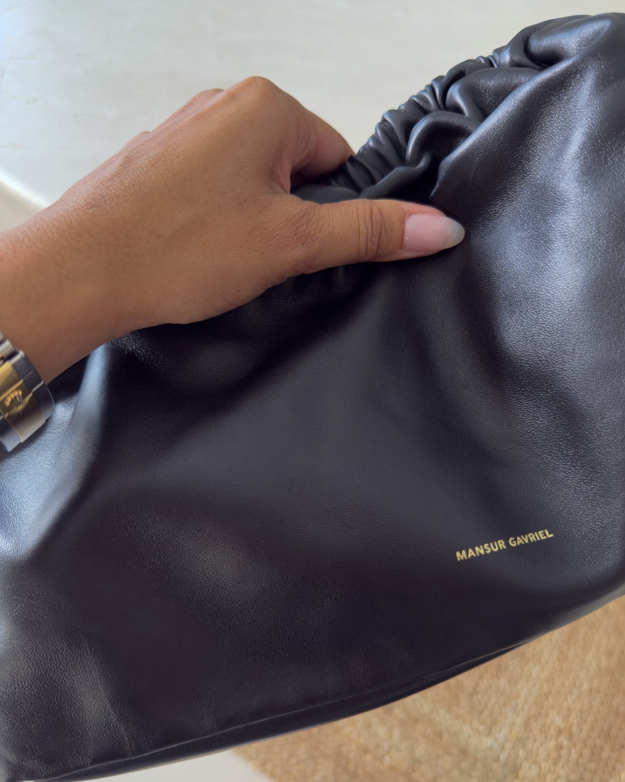Hand resting on the black lambskin of the Mansur Gavriel Oversized Cloud beside its gold logo stamp - Luxe Digital