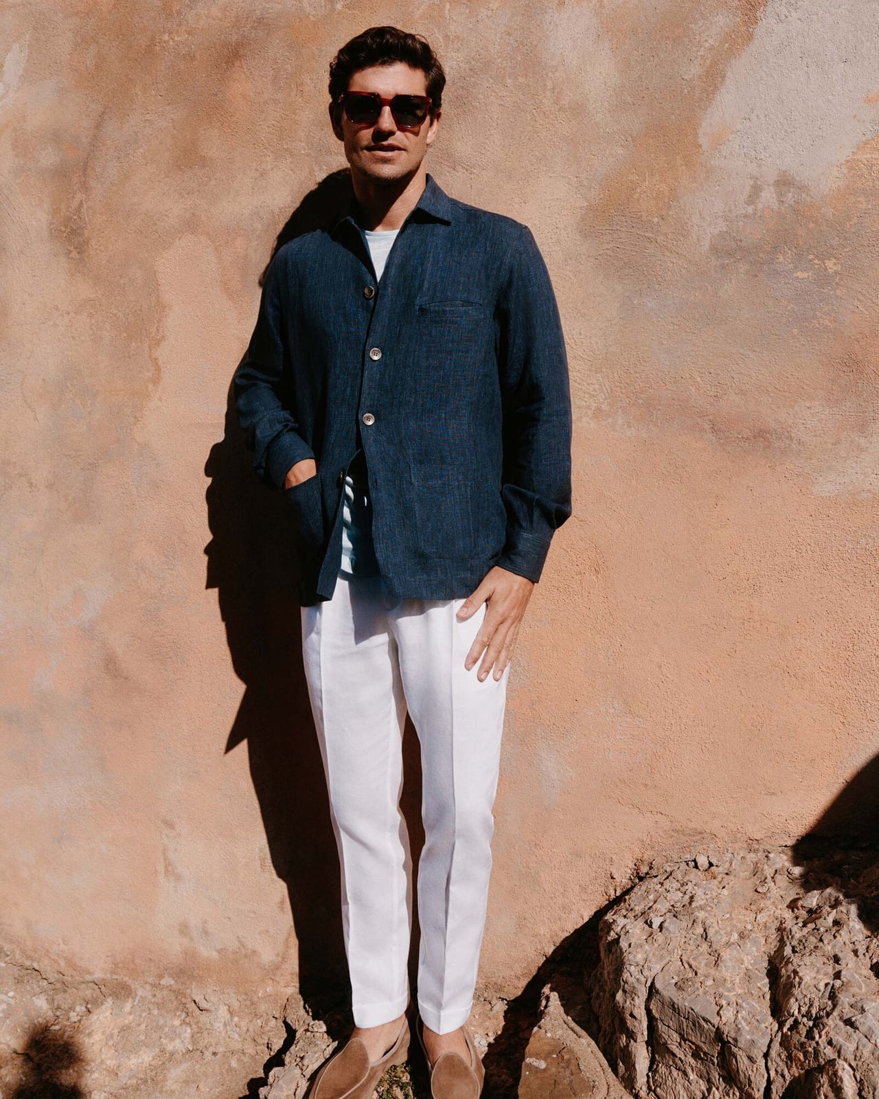 Man in a navy linen jacket, white T-shirt, white trousers, suede loafers and sunglasses against a terracotta wall