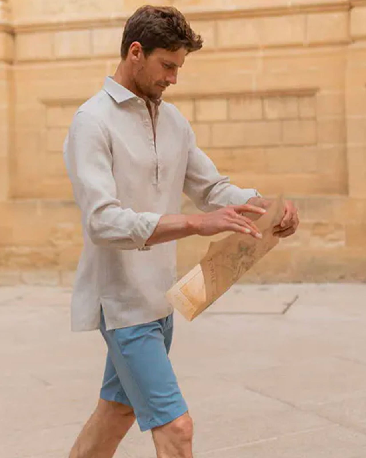 Man in a beige linen popover shirt and blue shorts reading a map on a sunny street