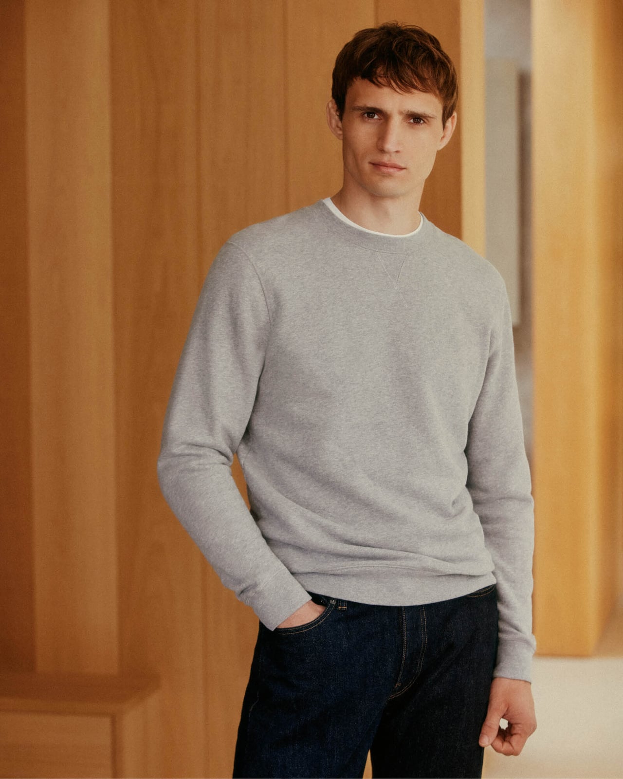 Man in a grey crew-neck sweater and dark jeans leaning against a wooden wall