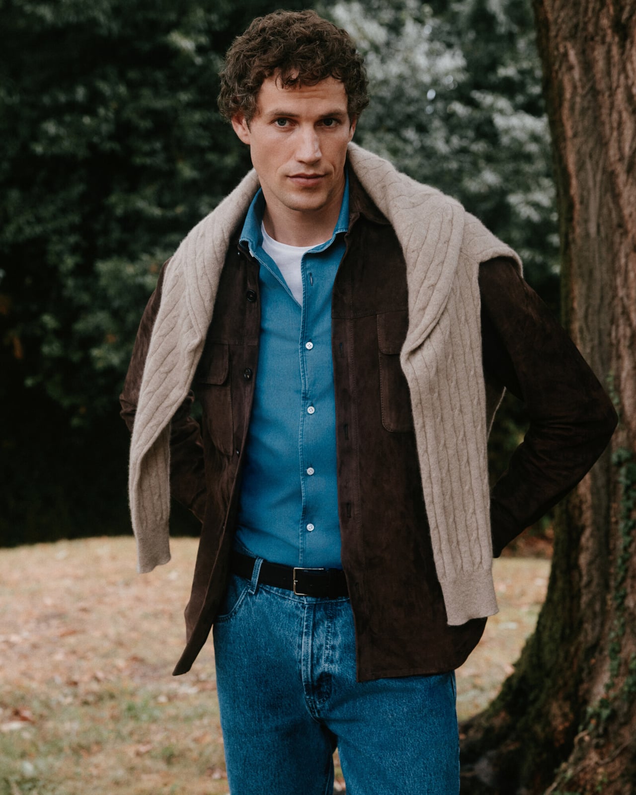 Man in a brown suede overshirt, blue shirt and jeans with a cable-knit sweater over his shoulders