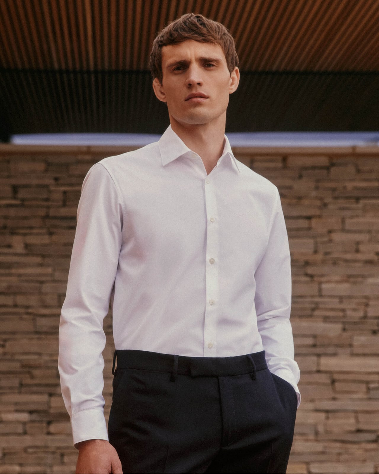 Man in a white shirt and dark trousers standing in front of a stone wall