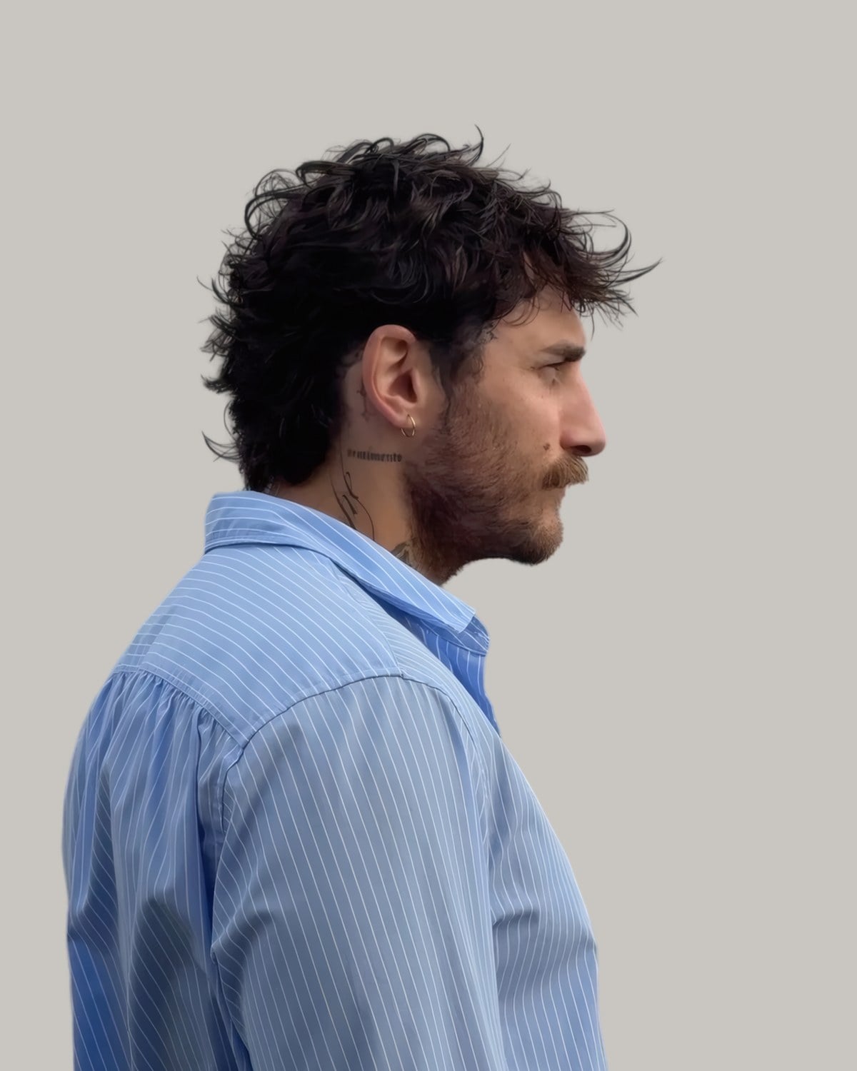 Bearded man with a messy dark curly mullet, seen in profile, in a striped shirt