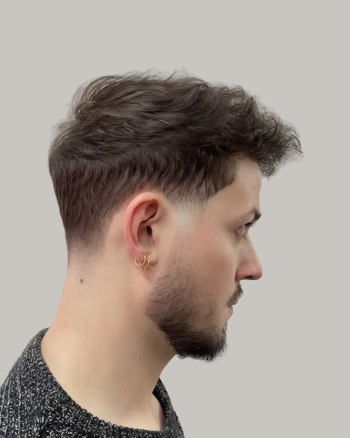Man with a stubble beard, a gold hoop earring and a messy textured top, seen in profile