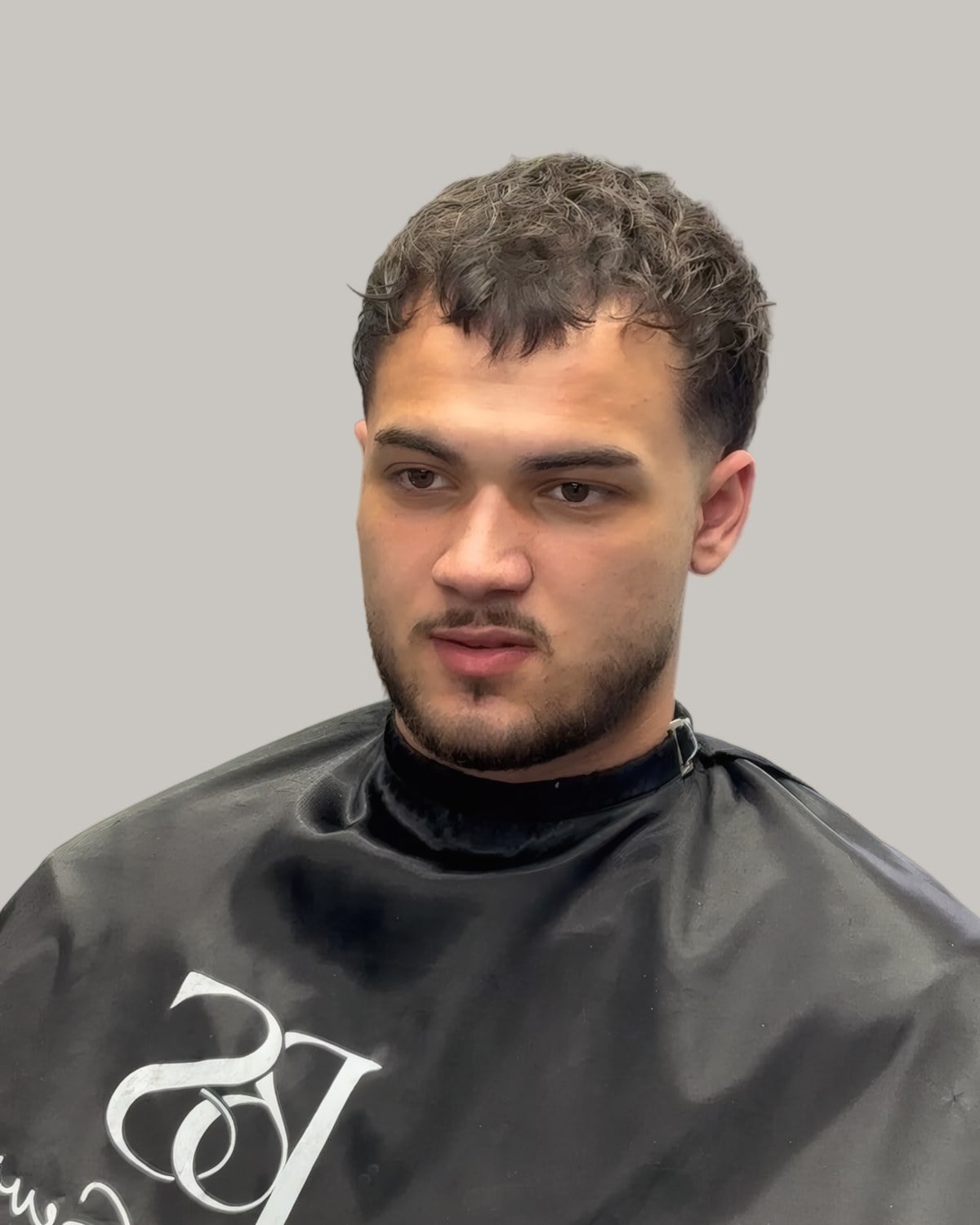 Man with a short Caesar cut and a trimmed beard, in a barber's cape, seen from the front