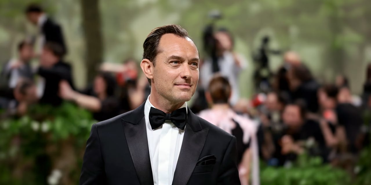 Man in a black tuxedo and bow tie with short hair slicked back from a receding hairline, at an outdoor event