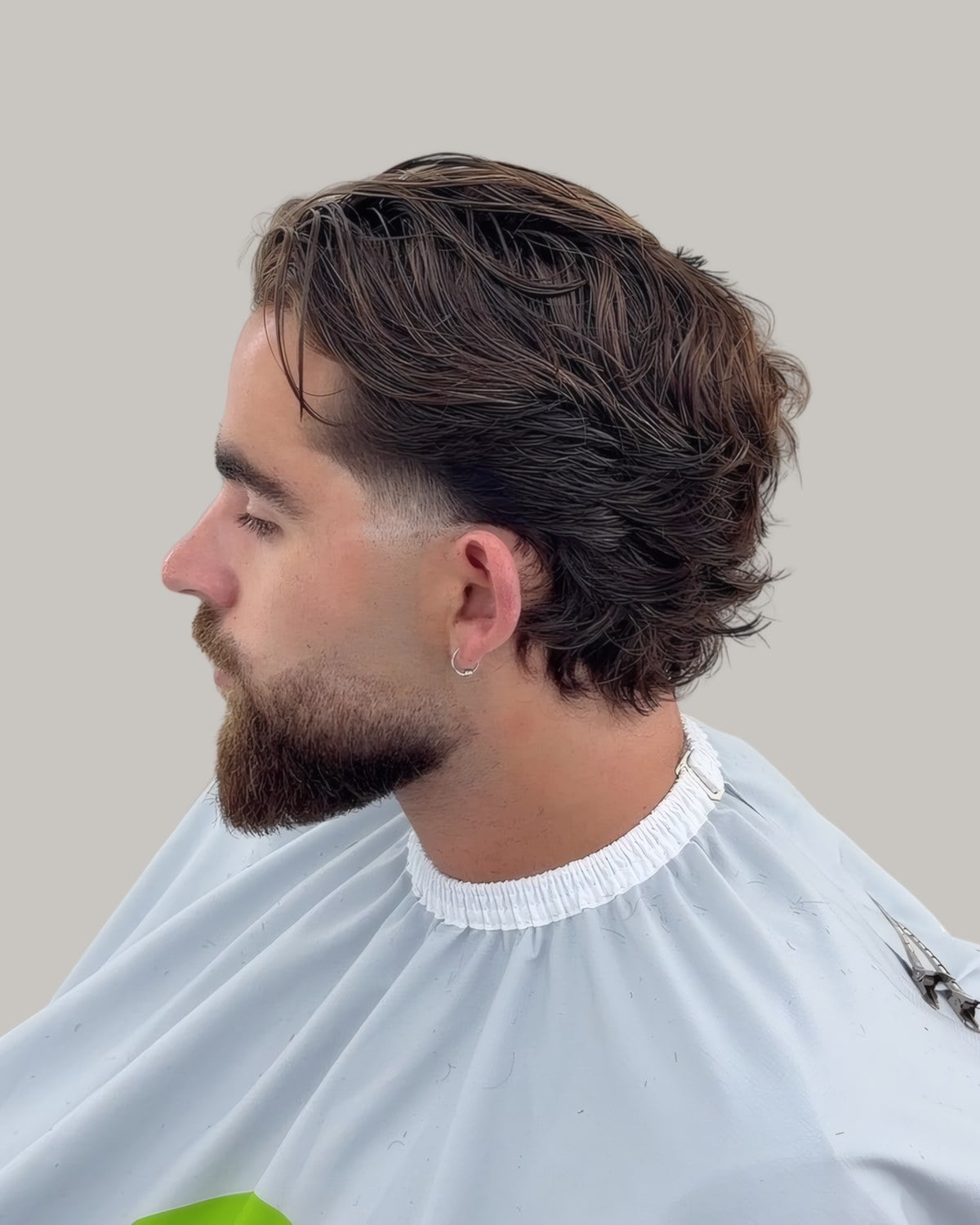 Bearded man with medium-length hair swept back, in a barber's cape, seen in profile