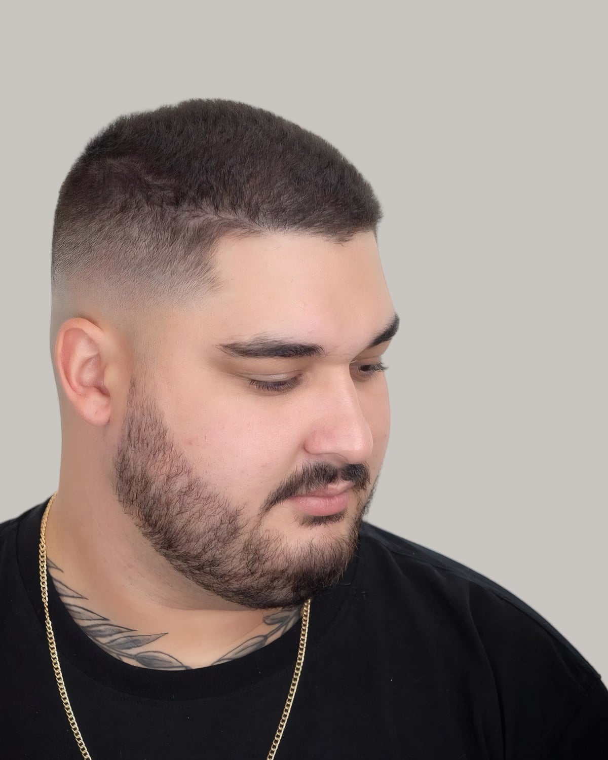 Bearded man with a short buzz cut and a skin fade, wearing a gold chain, looking down