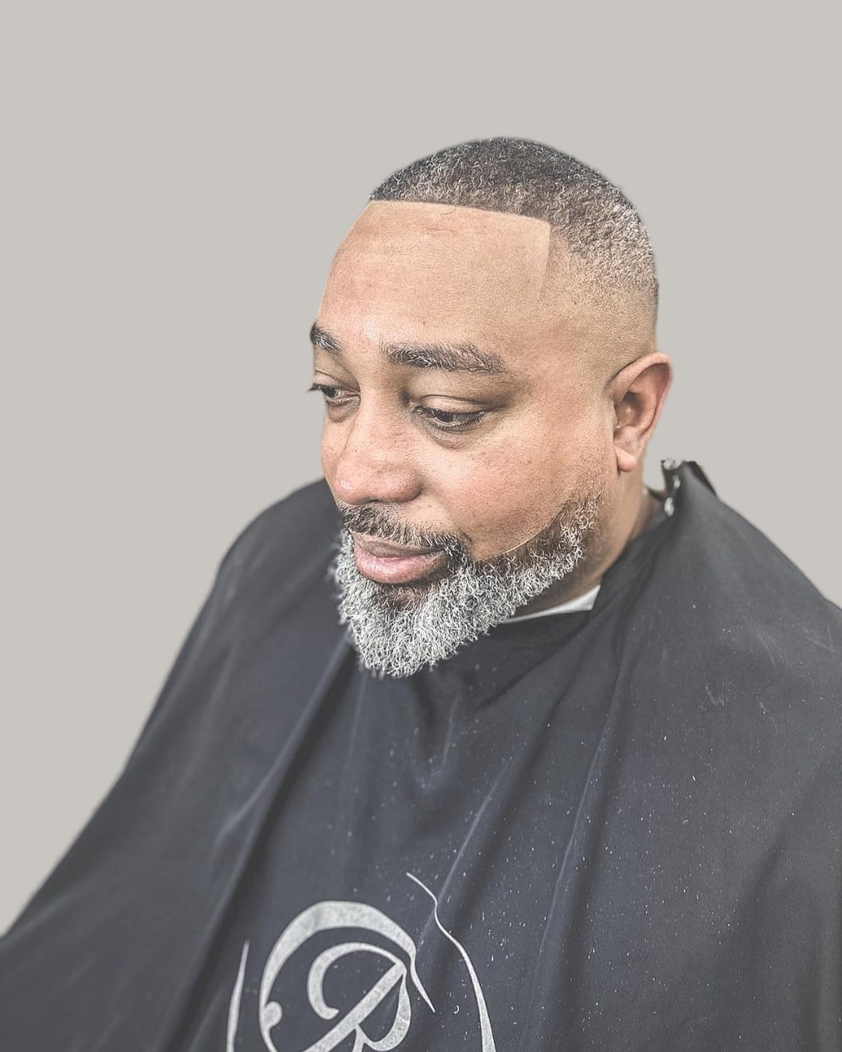 Older man with a short crop, a high skin fade, a squared line-up and a short gray beard, in a barber's cape, looking down