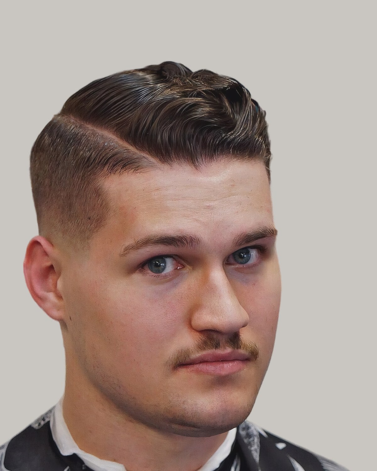 Man with a wavy comb over, a shaved hard part and a mustache, in a printed barber's cape