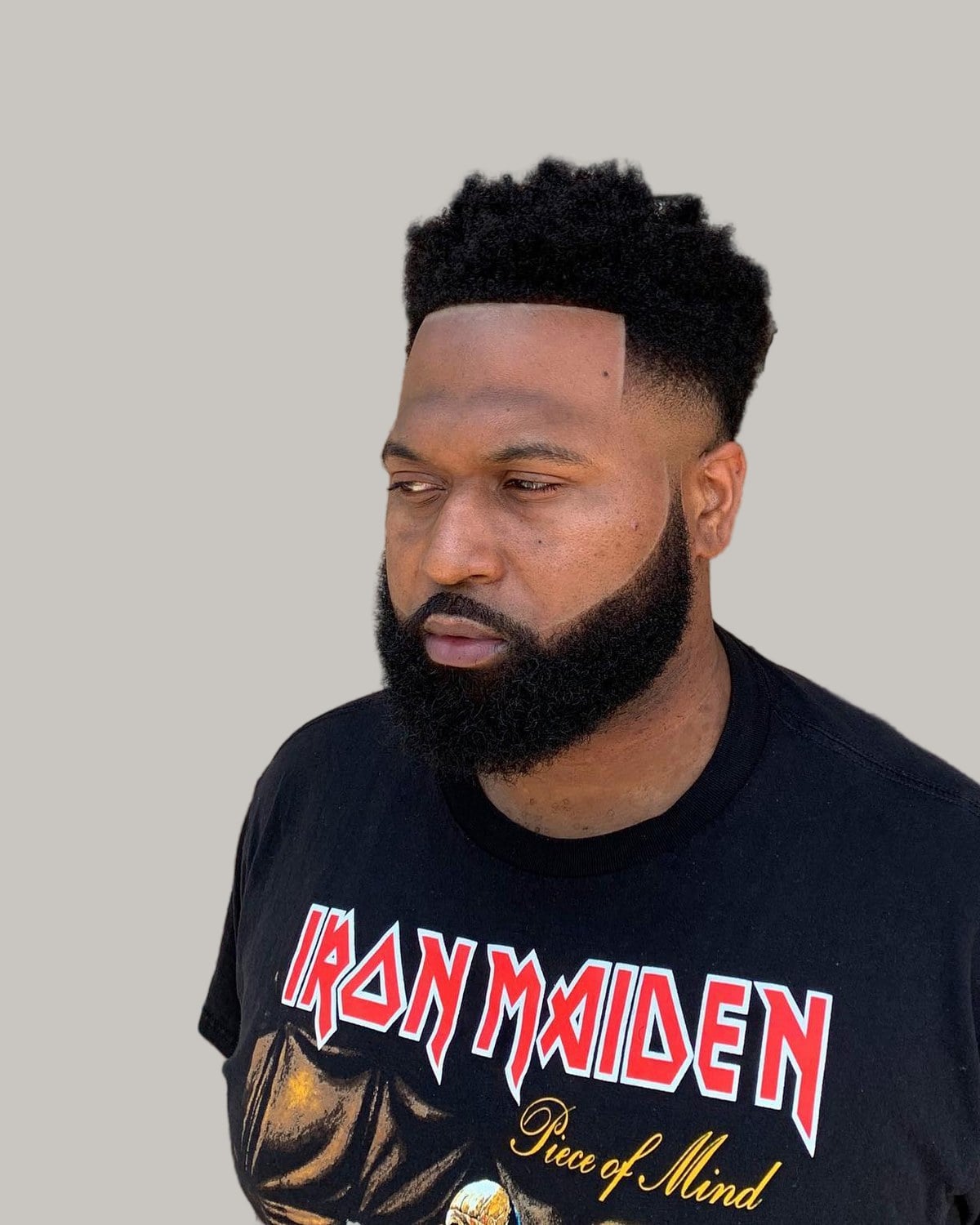 Bearded man with a short, curly quiff, a high fade and a sharp line-up, in a black band T-shirt, looking to the side