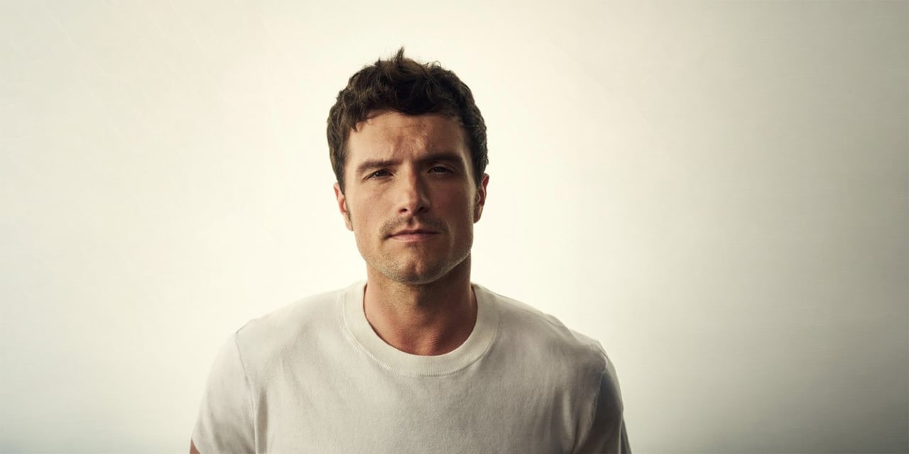 Man with short, tousled dark-brown hair and light stubble, in a white T-shirt against a pale backdrop