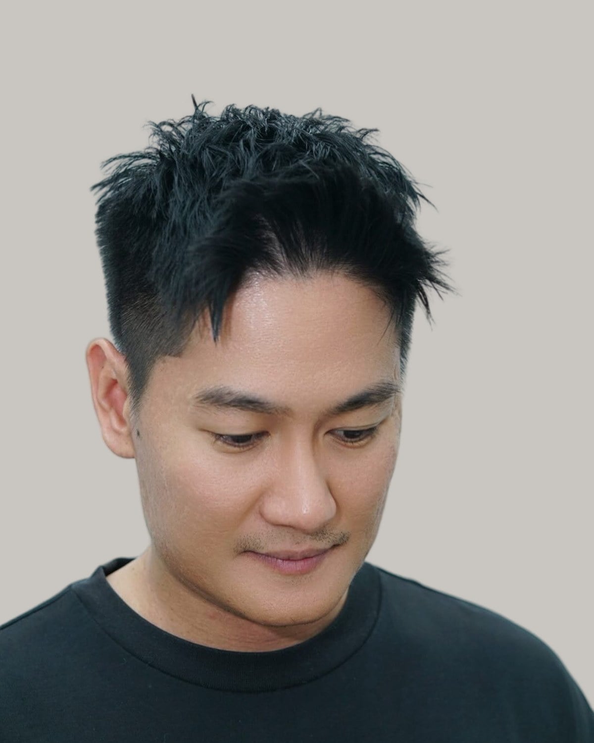 Man with a messy, spiky textured crop and short tapered sides, in a black sweatshirt, looking down