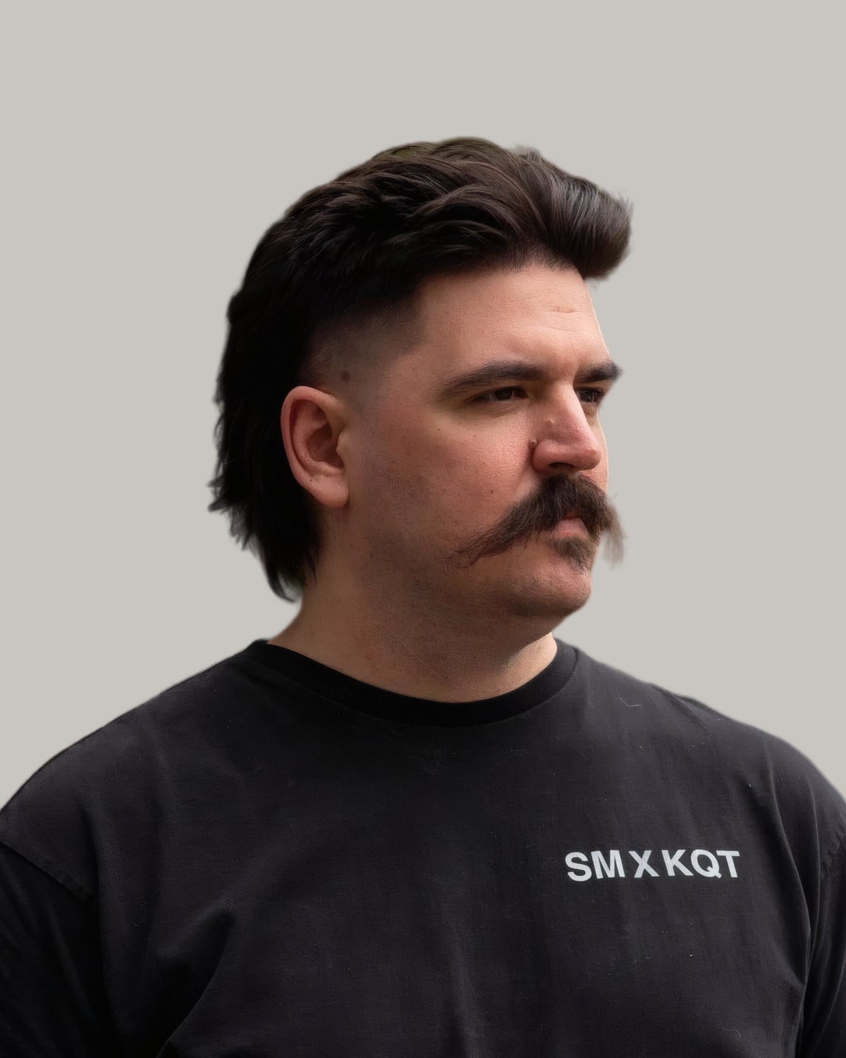 Man with a dark mullet, faded sides and a thick mustache, in a black T-shirt, looking to the side