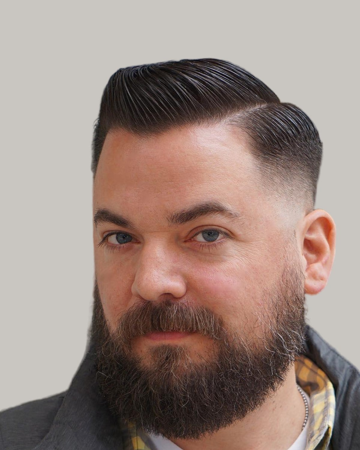 Man with a slicked side part, a skin fade and a long, full beard, in a gray jacket over a checked shirt