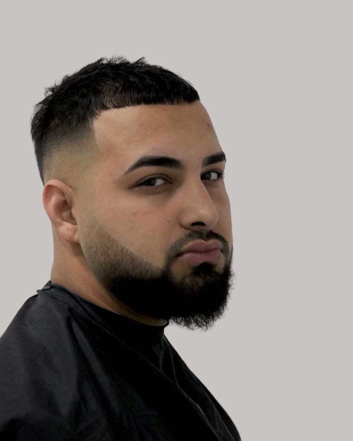 Bearded man with a short top, a skin fade and a pointed beard, in a black barber's cape, seen in three-quarter view