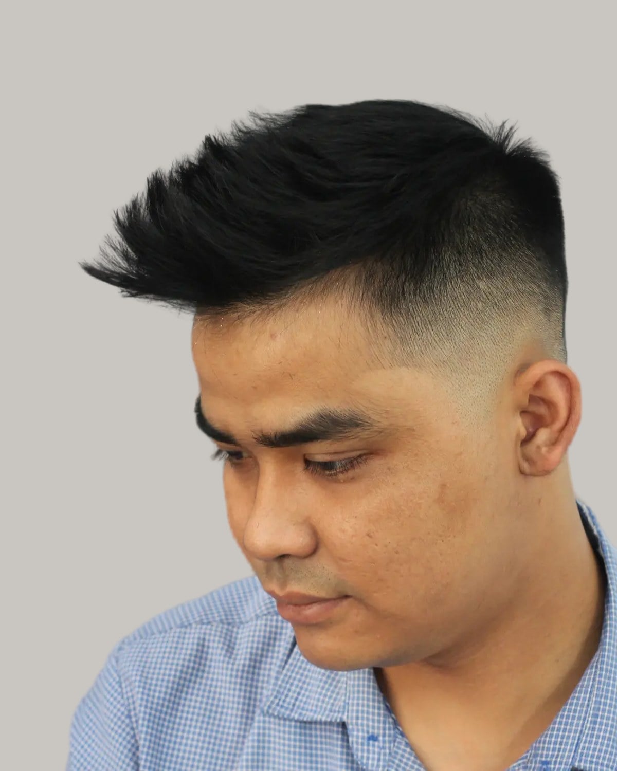 Man with a spiky black quiff and a high skin fade, in a blue checked shirt, looking down to the side