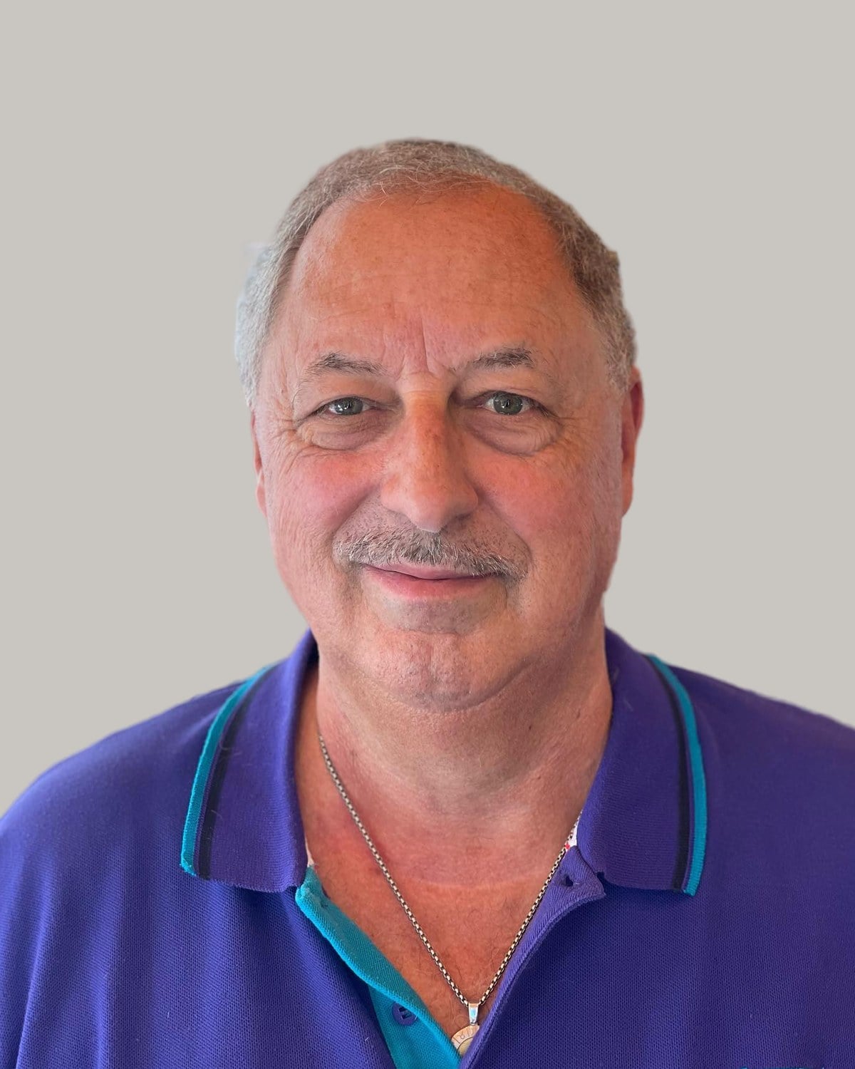 Smiling older man with short, thinning gray hair and a mustache, in a purple polo shirt