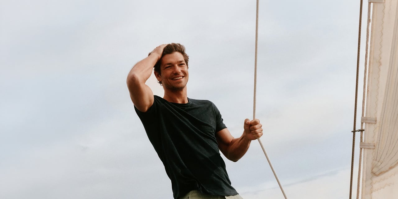 Smiling man in a dark T-shirt running a hand through his short brown hair, holding a rope on a sailboat