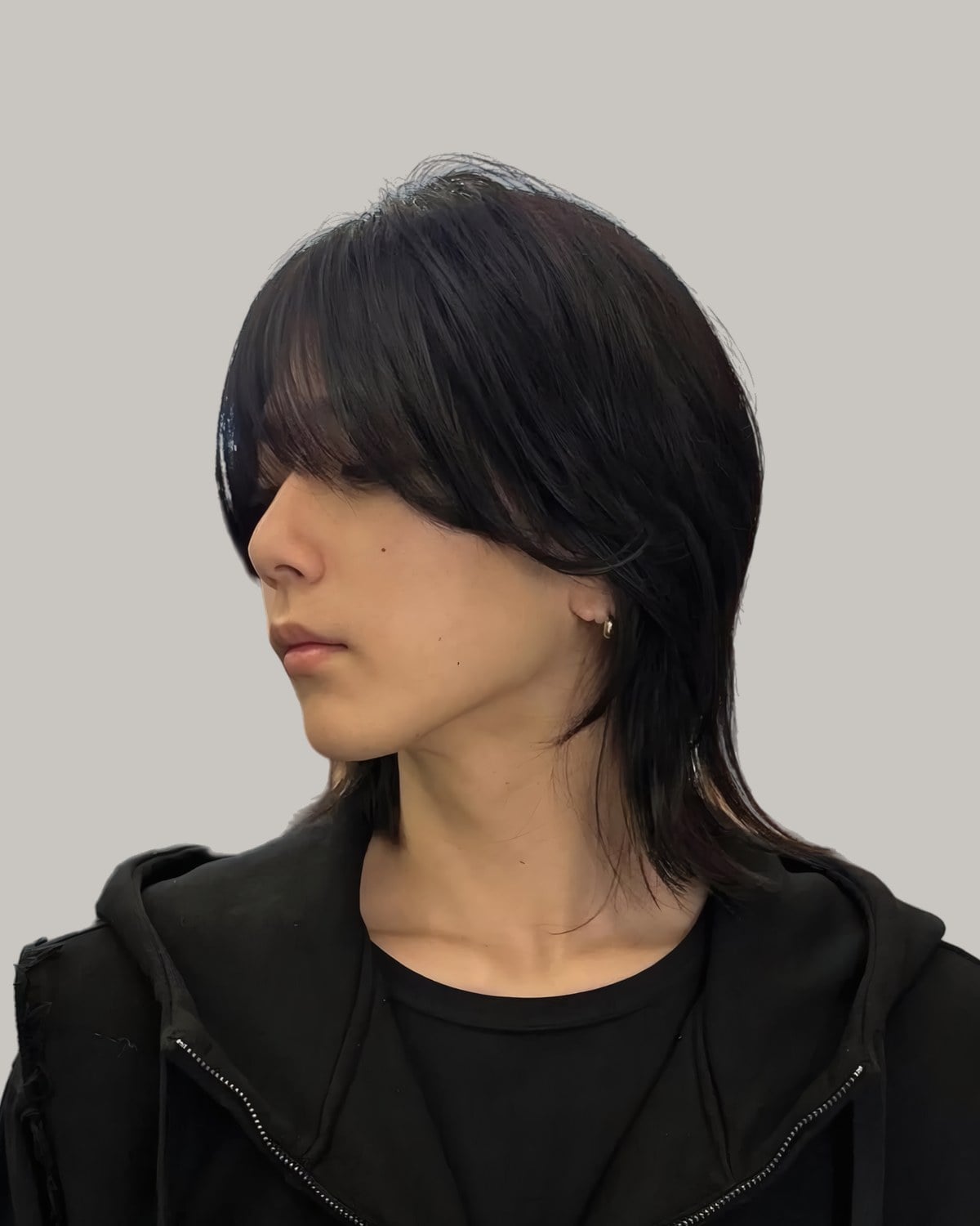 Young man in a black hoodie with long, layered black hair and a fringe falling over one eye