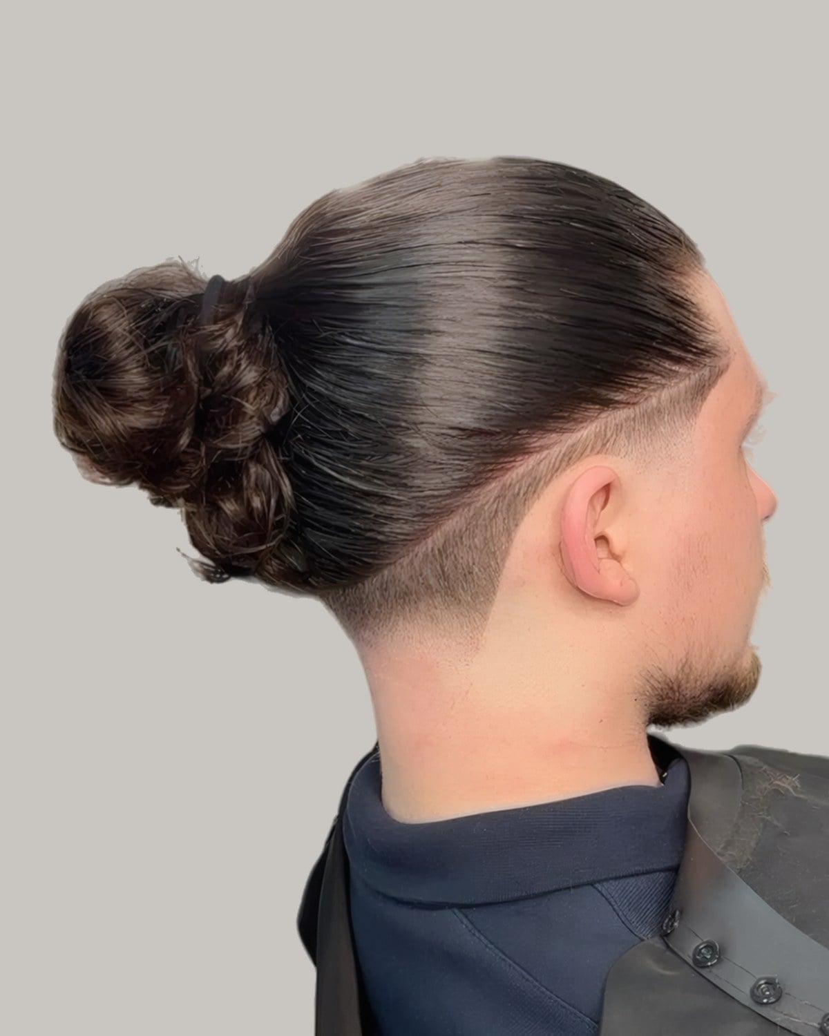 Man in a barber's cape with his long top tied in a bun over a faded undercut, seen from behind