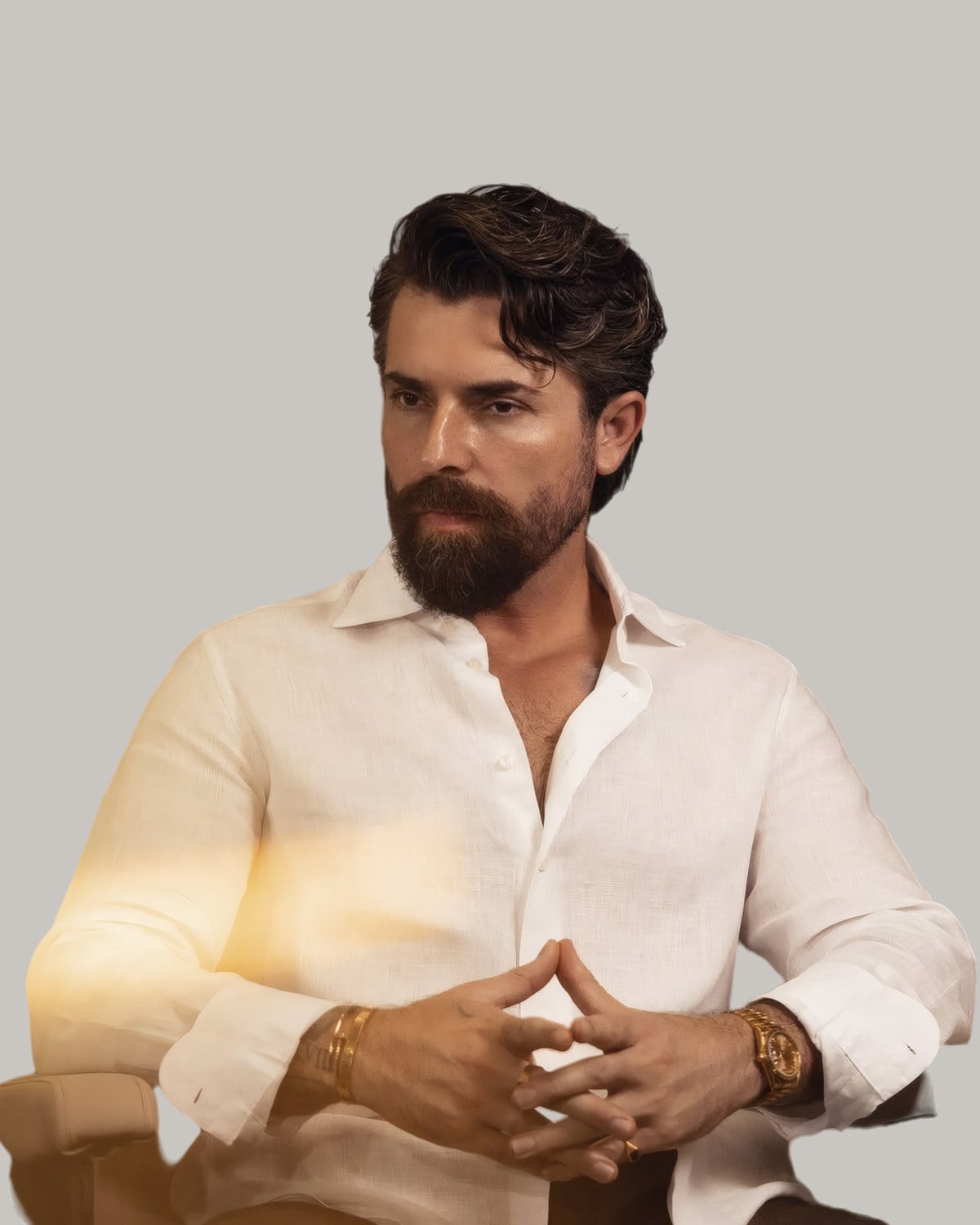 Bearded man in a white linen shirt with his wavy dark hair combed back, seated