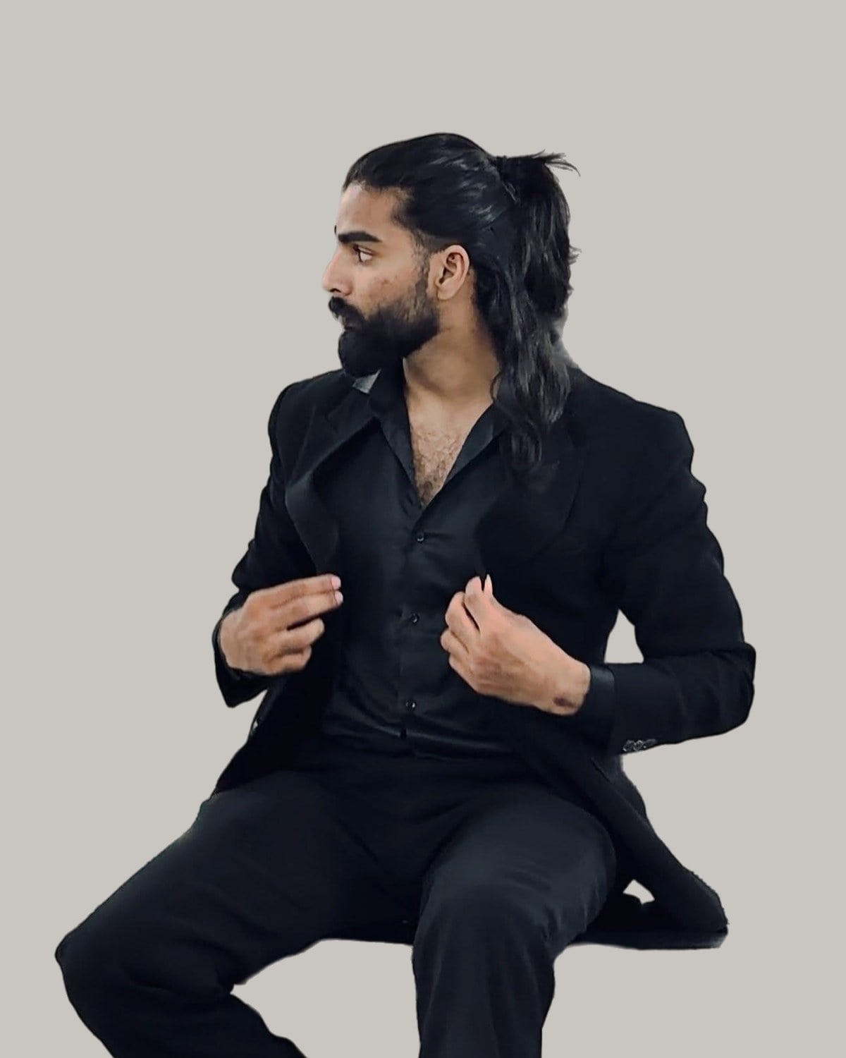 Bearded man in a black suit with his long hair swept back and tied, seated and looking to the side
