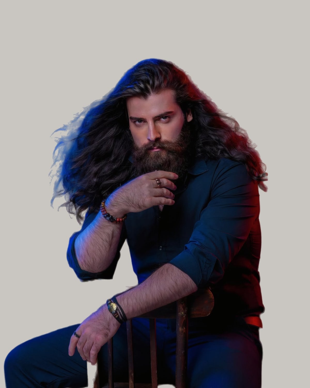 Bearded man with long, wavy dark hair, seated on a wooden chair with a hand at his beard