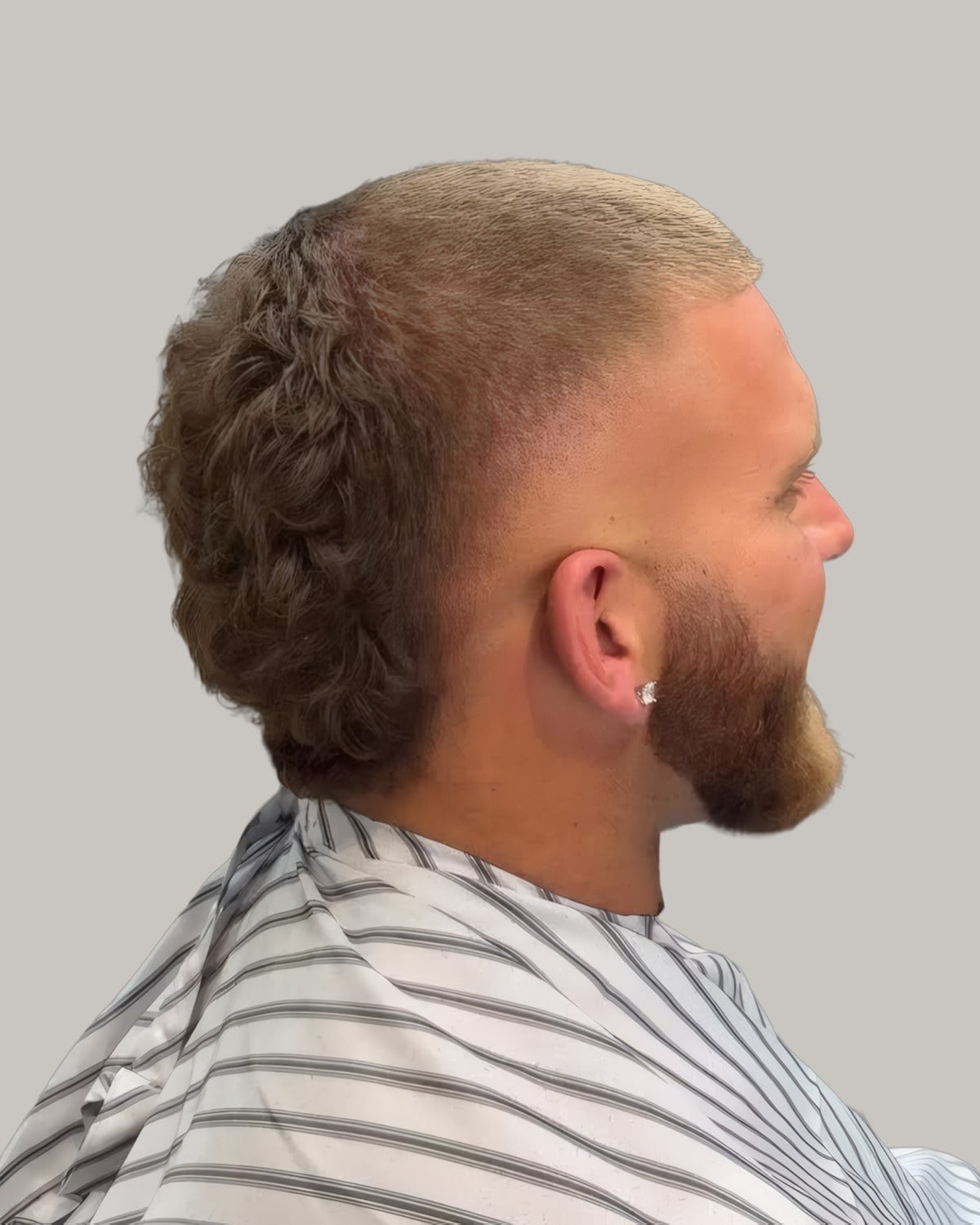Bearded man in a barber's cape with buzzed hair on top and longer hair at the back, seen from behind at an angle